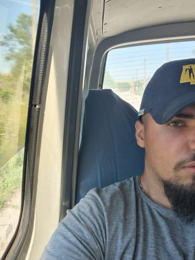 A close-up, self-portrait style photograph shows a man wearing a navy blue baseball cap with a yellow emblem and a grey t-shirt. He has a beard and a mustache and is looking towards the camera. He is seated inside a vehicle, likely a van or truck, with a blue seat visible behind him. The window of the vehicle is to his left, showing a blurred green landscape with trees and a dirt road outside. The scene appears to be set in a rural area, possibly Lomnitsa, Bulgaria, given the natural vegetation and the presence of utility poles and wires in the distance, visible through the rear window of the vehicle. The lighting suggests it is daytime.