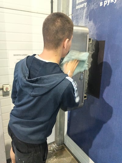 A young male, seen from the back and right side, is standing at a car wash station in Spassk-Dal'nij, Russia. He is wearing a dark blue hooded sweatshirt and dark shorts, and is wiping down a metal payment machine with a light blue cloth. The machine has a black rectangular panel on its right side. Behind him, a white corrugated wall is visible, with a sign that reads "ВНИМАНИЕ!" (ATTENTION!) and "ТЕРМИНАЛ" (TERMINAL) in Russian. To the right of the payment machine is a textured blue wall with faint white markings. The floor appears to be wet and dirty. The lighting is artificial and suggests an indoor or covered environment.