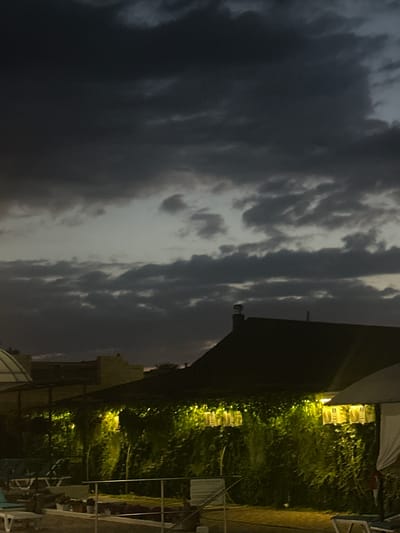 The image depicts an outdoor scene at dusk, possibly a hotel or resort area, in Yaroslavskaya, Russia. The sky is filled with dark, textured clouds, suggesting an overcast or potentially stormy evening. In the foreground, a building with a dark roof is visible, partially obscured by lush green foliage. String lights are illuminating the greenery, casting a warm, yellow glow. Several outdoor seating areas with lounge chairs are present, some appearing to be near a pool area, which is hinted at by the presence of loungers and what might be the edge of a structure. The overall atmosphere is dim and twilight, with artificial lighting providing a contrast to the natural light of the fading day. There are no people clearly visible in the image.