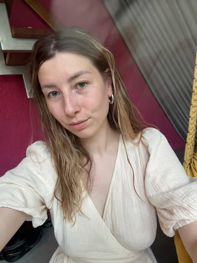 A young woman with light brown hair and blue eyes is posing for a selfie. She is wearing a light-colored blouse. The background includes a burgundy surface, a gray striped surface, and a gold rope. The location in the city of Чёрное, Russia is not discernible from the provided image.