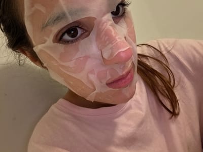 A human female is depicted in a close-up, slightly angled portrait, taken indoors in Plasencia, Spain. Her face is covered with a white, translucent sheet mask, which has apertures for her dark brown eyes, nostrils, and pink lips. Dark brown hair is visible, falling over her right shoulder. She is wearing a light pink, possibly cotton, crew-neck garment. The immediate background consists of a plain, light-colored wall. The subject's gaze is directed towards the camera.