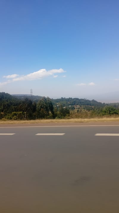 A sunny landscape presents a wide asphalt road with white dividing lines dominating the foreground. Beyond the road, a stretch of green hills and trees forms the midground, punctuated by a distant utility pole. Small buildings dot the hillside. Above, a clear blue sky, sparsely populated with a few white clouds, completes the scene. The image exhibits a clear, bright, and somewhat hazy atmosphere.