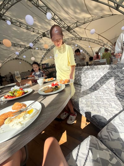 The image depicts an outdoor dining scene under a white tent structure, possibly in Morozovo, Russia, characterized by its open-air ambiance and decorative elements like hanging white orbs. In the foreground, a wooden table is laden with breakfast dishes, including a salad, a croissant with a fried egg, and other food items on plates. A glass of champagne is also visible. A young child wearing a dark cap and a yellow shirt is standing at the edge of the table, looking down at the food. Further back, other individuals are seen near a buffet area, suggesting a communal meal or event. The seating arrangement includes a patterned sofa with geometric designs. The overall scene suggests a relaxed, possibly resort-like atmosphere during daylight hours, with sunlight casting shadows on the floor and furniture.