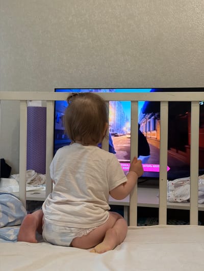 A young child, viewed from behind, sits in a crib facing a television screen displaying a city scene. The child wears a white t-shirt and a diaper. The crib has white wooden bars. Behind the crib, a shelf unit contains folded items and what appears to be a textured purple pad. The wall behind the crib has a textured, light-colored wallpaper. The image is taken indoors, likely in a bedroom or nursery. The location is identified as Shikhovo, Russia, though no specific visual cues in the image confirm this.