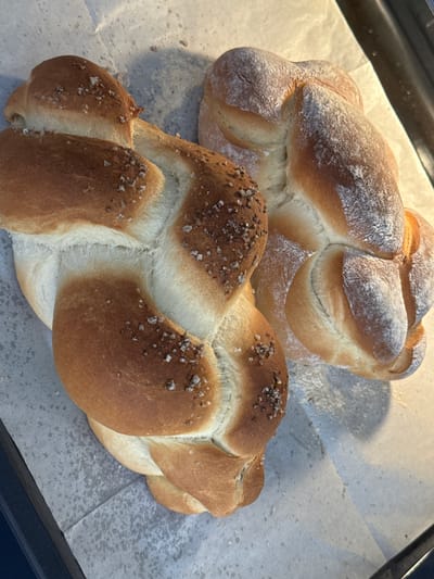 Two braided loaves of bread, topped with coarse salt and what appears to be a dusting of flour, are positioned on a baking sheet lined with parchment paper. The loaf in the foreground is on the left, and the second loaf is slightly behind and to the right. The lighting creates shadows that emphasize the texture and shape of the bread. The background consists of a textured surface, possibly a countertop or baking surface, with a dark edge of the baking sheet visible on the bottom left. There are no people or animals present in the image, and the scene is devoid of any contextual information indicating a specific location within Ariel, Palestinian Territories or any other city.