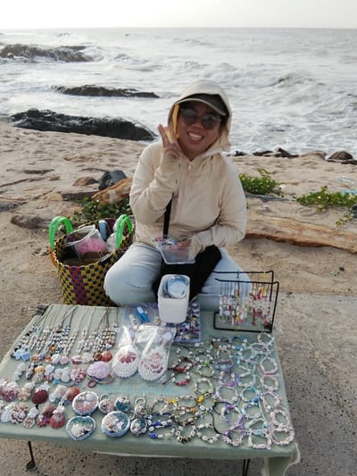 A woman is sitting on a sandy beach in Phu Thuy, Vietnam, with the ocean in the background. She is wearing a hooded jacket, sunglasses, and a cap, and is smiling at the camera while holding up two fingers in a peace sign. In front of her is a table displaying an assortment of handmade shell jewelry, including necklaces, bracelets, and pendants. Behind her, a woven basket filled with supplies and other items is visible, along with a black display rack for jewelry. The beach is covered in sand and scattered with rocks and some sparse vegetation. The waves are visible in the ocean, and the sky is overcast.