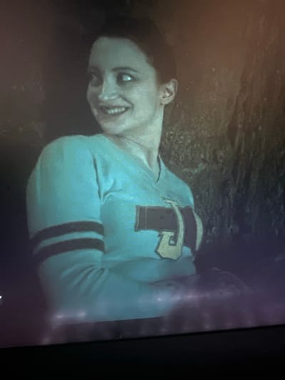 A close-up, dimly lit, medium shot shows a young woman with fair skin and dark hair. She is wearing a light blue, long-sleeved shirt with dark stripes around the left sleeve and a large, yellow letter "J" outlined in dark trim on the front. She is looking over her right shoulder with a wide, toothy smile and her eyes are also looking to the right. Her left arm is bent, with her hand resting on her chest. She is positioned in front of a textured, dark background that appears to be rock or earth. The image's lighting is poor, with noticeable artifacts and a soft focus. There is no indication of the location in the city of Vancouver, United States of America.