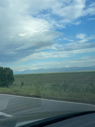 The image depicts a view from a vehicle, likely a car, driving along a road near Vodenichane, Bulgaria. The sky is predominantly blue with layered white clouds. A field extends to the right, punctuated by distant fence posts and hints of yellow wildflowers. Beyond the field are indistinct mountains. A large green tree is visible in the lower part of the frame. Part of the vehicle's interior and window is visible on the right edge. There are no people visible in the image.