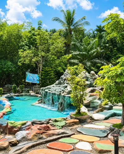 The image depicts a vibrant outdoor swimming pool area located in the city of Eampat Kwalamencirim, Indonesia. The pool is designed with artificial rock formations and a small waterfall, filled with clear turquoise water. Surrounding the pool is a colorful pathway made of large, oval-shaped stepping stones in various hues, laid over a bed of small grey pebbles. Lush green tropical vegetation, including palm trees and dense foliage, frames the scene under a bright blue sky with scattered white clouds. A banner with text that appears to say "WISATA TELAGA BAHARI" is visible on the left side of the pool, suggesting it is a tourist attraction. No people are present in the image. The overall environment suggests a recreational water park or resort setting.