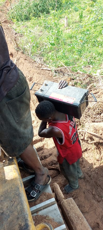 The image depicts a scene of manual labor in an outdoor, unpaved environment, likely during daytime given the bright natural light. The primary subject is a young child, possibly a boy, seen from the back and side. He is wearing a red sleeveless basketball jersey with "JORDAN" and the number "23" clearly printed in white on the back. The child is carrying a large, heavy-looking black battery on his head and right shoulder, steadying it with both hands. The battery features visible terminals and wire handles, and a red label with white, indistinct text is attached to its side.

Partially visible on the left side of the frame is an adult, whose legs and lower torso are shown. The adult is wearing dark shorts or trousers and black sandals with buckles, and is standing near a large, yellow piece of equipment, possibly machinery, which is also covered in dirt.

The ground is reddish-brown dirt, with a distinct trench or ditch running from the foreground towards the background. Beyond the immediate area of activity, lush green foliage and bushes are visible, indicating a rural or undeveloped setting. A wooden post or stake lies on the dirt near the adult's feet. The context provided indicates the location is Otunade, Nigeria.