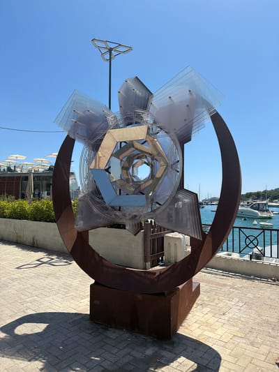The image shows a large, abstract sculpture with a rusted metal base and a circular frame. Inside the frame is a complex structure made of transparent plastic and mirrored geometric shapes. The sculpture is located on a paved walkway next to a body of water, likely the Sliema Creek in Gżira, Malta, as suggested by the presence of boats and the distinctive waterfront architecture in the background. The sculpture casts a long shadow on the ground, indicating it is a sunny day. In the background, there is a building with a rooftop terrace occupied by people under umbrellas. The water is calm and several boats are docked or anchored. The sky is clear and blue.