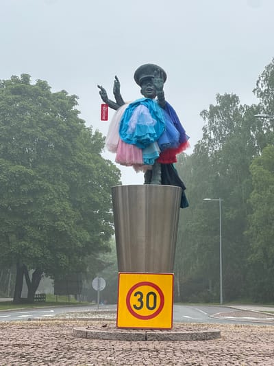 A bronze-colored statue, titled "The General's Good Day" (Kenraalin hyvä päivä) by Pekka Kauhanen, is centrally featured in Tapiola, Espoo, Finland. The sculpture depicts a male figure, typically identified as Marshal C.G.E. Mannerheim, wearing a dark cap. The figure's left arm (viewer's right) is raised with an open palm, while the right arm (viewer's left) is also raised with fingers pointing upwards. The statue is adorned with multiple layers of ruffled fabric skirts or tutus in various colors, including light blue, dark blue, pink, red, and dark teal. A red rectangular tag with white text, reading "SCHÖN!" (German for "beautiful" or "nice!"), hangs from a chain on the figure's raised right hand.

The statue stands on a tall, cylindrical pedestal made of brushed metal, resembling a large cup. This pedestal is situated on a circular base composed of reddish-brown cobblestones. Immediately in front of the pedestal, a yellow square sign with a red circular border and the number "30" in black is displayed, indicating a speed limit.

The background consists of a dense canopy of lush green deciduous trees, extending across the frame under an overcast, light grey sky. A paved road, featuring white dashed lines and arrows, curves around the base, indicating a roundabout configuration. Two streetlights with grey poles are visible against the backdrop of trees. The overall atmospheric condition is hazy, suggesting fog or a very light mist.