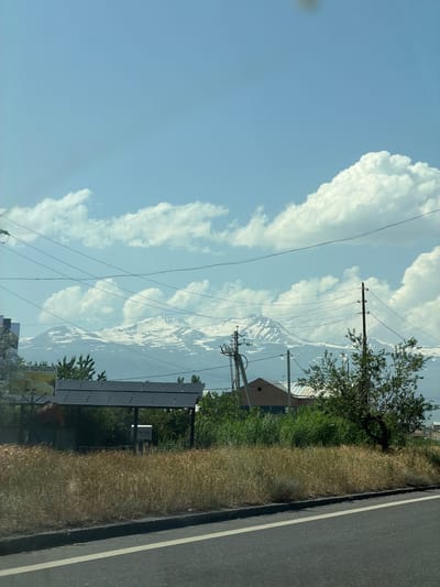 The image shows a view of Mount Ararat, a snow-capped mountain range, visible in the distance under a bright blue sky with scattered white clouds. In the foreground, there is a road with a white line marking the lane. Beside the road, there is dry grass, a small building with a distinctive brown roof, and some green trees. Power lines crisscross the frame, supported by wooden poles. To the left of the frame, a structure with solar panels is visible, with some signage that appears to be in Armenian. The overall scene suggests a semi-rural or suburban environment on the outskirts of the city of Proshyan, Armenia, with the iconic Mount Ararat serving as a prominent backdrop. There are no people or animals clearly visible in the image.