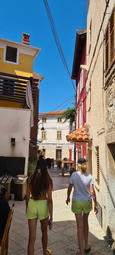 The image depicts two women walking down a sun-drenched street in a European coastal town, likely in Općina Fažana, Croatia. The scene is captured from a slightly elevated perspective, looking down the narrow, paved street lined with buildings of various colors and architectural styles.
The first woman, closer to the foreground and facing away from the camera, has long, dark, wavy hair and wears a black top and bright yellow-green shorts. She is holding a phone in her right hand. The second woman, slightly further ahead and also facing away, has blonde hair tied back in a ponytail and wears a white t-shirt and similar yellow-green shorts. Her left arm is extended slightly, as if gesturing or balancing. Both women are wearing sandals.
The buildings showcase a mix of textures and colors, from rough-hewn stone to smooth plastered walls in shades of red, yellow, and white. Wooden shutters are visible on many of the windows, suggesting a Mediterranean or Adriatic architectural influence. The clear blue sky indicates fair weather, and the strong sunlight casts distinct shadows on the cobblestone street, suggesting it is midday or early afternoon.
Objects in the scene include outdoor seating and tables of a cafe or restaurant, a terracotta-tiled roof detail on one of the buildings, and power lines overhead. Green foliage is also visible, climbing up some of the walls, adding a touch of nature to the urban environment. The overall atmosphere appears relaxed and casual, with the women likely enjoying a stroll through the town. No legible text is visible in the image.
