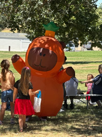 The image depicts an outdoor scene in Elverta, United States, on a sunny day with dappled light filtering through trees. In the foreground, an inflatable character, appearing to be an orange creature with a brown face and green headpiece, stands prominently. To the left of the character, two young girls are visible from behind. The girl on the left wears a tan t-shirt and denim shorts, while the girl on the right wears a black t-shirt, a red skirt adorned with small white hearts, and sandals. She is holding a white tote bag with blue handles, which has a black line drawing on it.

Further back, a group of people are gathered near a picnic table. A woman in a dark blue jacket with white stripes is seated on the right side of the table, facing left. In front of her, a young girl with blonde hair in pigtails wears a pink shirt and a red skirt. She is reaching out her hand towards the woman. Another person is seated at the table, wearing a white t-shirt. Behind the table, two adults and a child are partially visible. The background includes a grassy field, a building with white siding and a gray roof, and parked cars in the distance. The overall atmosphere suggests a casual outdoor event or gathering.