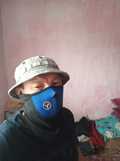 A person wearing a digital camouflage bucket hat and a blue and black face mask is pictured indoors. The person is looking directly at the camera. The background shows a pink, textured wall and some piled-up fabric items. The person is wearing a dark blue t-shirt. The image appears to be a selfie taken in an indoor setting. There is no specific indication of the city of Kojomkul, Kyrgyzstan, in the image.