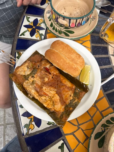 A white plate holds a tamale wrapped in a banana leaf, a small bread roll, and a wedge of lime. The tamale is covered in a reddish sauce. The plate sits on a colorful mosaic tile table. A cup and saucer are on the left side. A glass of beer is at the top left. A fork pierces the bottom of the tamale, and two people are visible in the image, one on the left and one on the right.