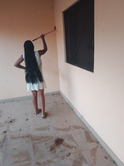 A young woman with long, dark braids stands on a tiled floor, holding a stick and seemingly cleaning the wall. She wears a light-colored dress and sandals. The floor is stained and dirty with debris. A window with a dark screen is visible on the right wall. The walls are painted in a muted, warm color.