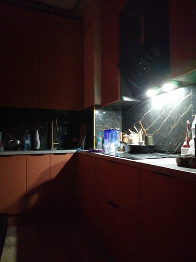 The image shows a kitchen counter with cabinets below and a backsplash with a marble-like pattern. On the counter, there are several objects including bottles of liquid, a metal bowl, and some kitchen utensils. A faucet is visible above the sink area. The lighting in the scene is artificial and appears to be coming from under the cabinets. The cabinets are a dark reddish-brown color, and the countertop is a lighter gray. The overall impression is of a functional kitchen space. The city of Trubacheyevka, Russia, is mentioned but there are no visual cues in the image to confirm or deny this location. There are no people or visible actions taking place.