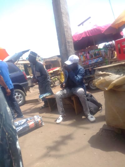 The scene is an outdoor commercial area in Mista Ali, Nigeria, under strong daylight. In the mid-right, a man sits on a wooden bench, wearing a blue jacket, light-colored jeans, white sneakers, and a white baseball cap pulled low; his right hand is near his mouth. A black backpack rests on the ground behind him. To his left, another man stands next to a dark blue vehicle, possibly a hatchback, with its tailgate open. This standing man wears a grey long-sleeved tunic with a light grey chest panel, dark trousers, and a patterned cap. A third individual, partially visible on the far left, wears a blue shirt. On the ground in the immediate foreground are several bundles of black plastic-wrapped merchandise with orange securing bands. A concrete pillar, marked "NIPP 32," stands centrally. Behind the seated man, a market stall with red and yellow canopies displays bottled and packaged items. Buildings are visible in the background, and the ground is unpaved earth.