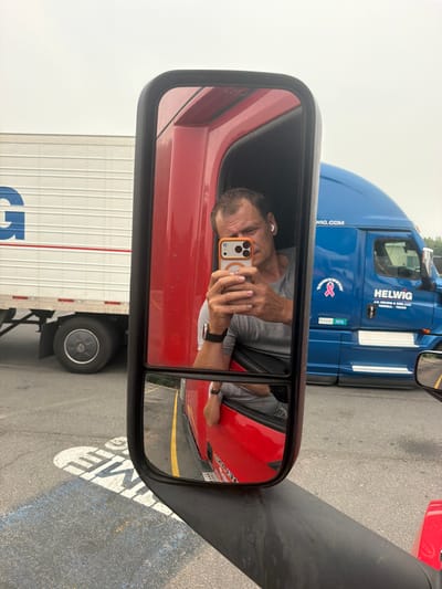 A Caucasian man in his 40s, with short brown hair and a grey t-shirt, is taking a mirror selfie in the driver's side mirror of a red semi-truck. He is wearing white wireless earbuds and a black watch. The truck has a red interior visible in the mirror. In the background, a blue semi-truck with "HELWI G" written on it and a pink ribbon logo is parked. To the left of the blue truck, a white semi-trailer with a large blue "G" logo is partially visible. The ground is asphalt with white and yellow painted lines. A parking lot marking with white letters "TIME" is visible in the lower left corner. The sky is overcast. The location is identified as Union, United States of America.