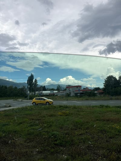 The image presents a ground-level view captured through the window of a vehicle, looking out onto a street scene in Pirdop, Bulgaria. The upper third of the frame shows a heavily overcast sky with uniform grey clouds, suggesting an impending or recent rain. A distinct horizontal line, representing the vehicle's window frame, separates this from the lower portion of the image. Below this line, the reflection in the window reveals patches of clearer blue sky interspersed with white cumulus clouds, indicating a different atmospheric condition or direction of light relative to the direct view.

In the foreground, a strip of unmaintained grassy land is visible, characterized by dry patches, various weeds, and scattered small yellow wildflowers. Detritus, including what appears to be plastic refuse, is sporadically present on the ground.

The midground features an asphalt road with white pedestrian crossing markings. To the left, a portion of a circular intersection or roundabout is discernible. A yellow Renault Clio hatchback, identifiable as a taxi by a roof-mounted sign, is depicted in motion, traveling from left to right across the road.

Beyond the road, a diverse landscape unfolds. On the left, several tall, slender deciduous trees stand prominently, flanked by other green foliage. Adjacent to these trees, a green street sign, partially visible, displays Cyrillic text including "ул. Септември" (Septemvri St.), a common street name in Bulgarian cities, confirming the location.

Further to the right, a complex of low-rise buildings with grey facades and distinctive red-trimmed roofs is present. One building prominently features a sign with the Cyrillic letters "УК" (UK). A blue square sign with a white triangular pictogram of a walking person (indicating a pedestrian crossing) is affixed to a pole near these structures. Utility poles and overhead lines traverse the scene.

The background is dominated by a distant, undulating mountain range, appearing muted in tone under the partially cloudy sky. No human figures are distinctly visible. The overall scene depicts a functional urban-natural interface on an overcast day.