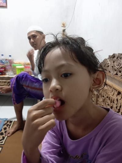 An indoor scene in Bulukandang, Indonesia, depicts two individuals. In the foreground, a young female, approximately 5-10 years old, with dark hair and dark eyes, wears a long-sleeved purple top and a gold-colored earring in her right ear. She is observed bringing a small, pink, spherical object to her open mouth with her right hand. To her right, a carved wooden furniture piece is partially visible. In the background, an adult male, wearing a white cap and a white long-sleeved shirt with purple trousers, sits barefoot on a low platform. He holds a piece of food in his raised right hand, with his mouth slightly open, looking to the viewer's left. Behind him, a white wall features a visible electrical outlet and wires. A small table near the male holds several plastic bottles and various plastic containers, some containing food or snacks. The floor is covered by a brown patterned rug.