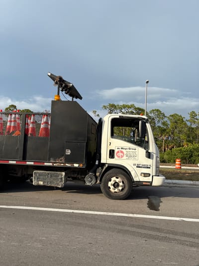 A white Isuzu truck with a black utility bed is parked on the right side of a two-lane road. The truck bed is loaded with orange and white traffic cones. A light is attached to the top of the truck bed. The truck has a "de Moya Group" logo on the passenger door. A US DOT number, 948028 FL, is visible on the truck's front fender. The background consists of green trees and a cloudy sky. An orange traffic barrel is visible on the side of the road. The scene is likely in Venice, Florida, United States.