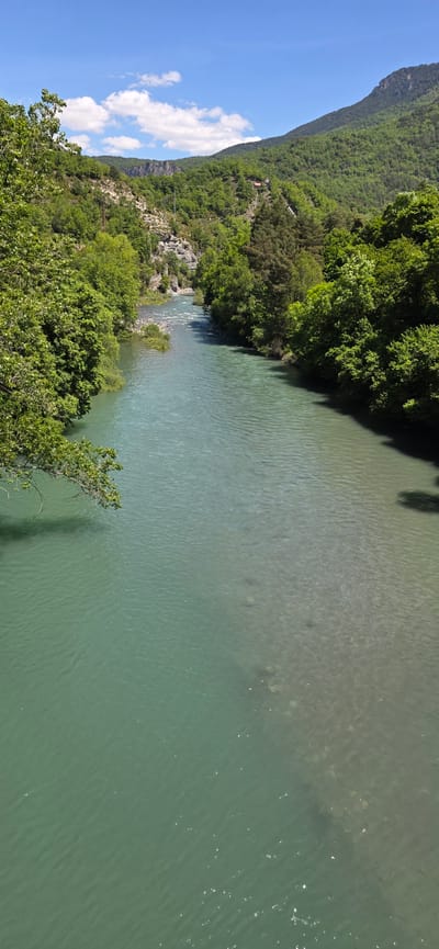 The image shows a river in Seira, Spain. The river is wide and the water is a greenish-blue color. The river is bordered by lush vegetation on both sides, with trees lining the banks. A mountain range is visible in the background. The sky is blue with some white clouds.