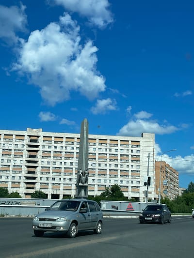The image captures a bright, daytime scene on a street in Kirovo-Chepetsk, Russia. The sky is a vibrant blue, dotted with fluffy white clouds. In the background, a large, multi-story residential building with a light-colored facade and rows of windows dominates the skyline. To the right of this building, another, older brick building is visible. In the foreground, a wide road is present with several cars. A silver station wagon is prominently featured, its license plate reading "E 987 HP 43." To its right, a dark-colored sedan is also driving on the road. Between the buildings and the road, there appears to be a monument or obelisk with sculptures at its base. The overall environment suggests an urban setting on a clear day.