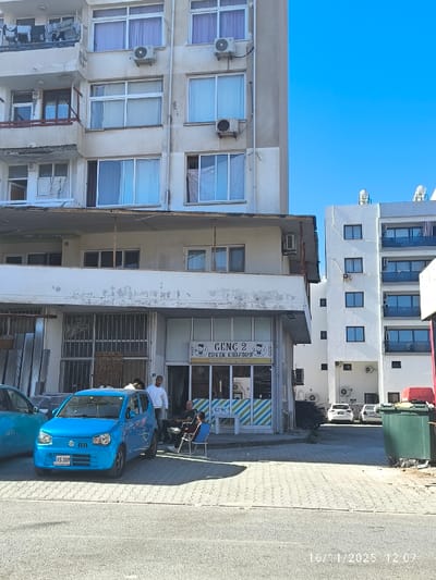This image depicts a street scene in Gonyeli, Northern Cyprus, on November 16, 2025, at 12:07 PM. The scene is dominated by a weathered, multi-story building on the left, featuring numerous windows with white frames and several wall-mounted air conditioning units. Clothes are visible drying on a upper-floor balcony. At the ground level of this building, the business "GENC 2 ERKEK KUAFORU" (Men's Barbershop) is visible, characterized by a glass storefront with a diagonally striped blue, green, and white panel.

In front of the barbershop, three adult males are present: one stands near the entrance in a white long-sleeved shirt, while two others are seated on foldable chairs outside, facing each other. A light blue compact car, possibly a Suzuki Alto, with license plate "VS 209," is parked on the paved street directly in front of the barbershop. The front of a second turquoise vehicle is partially visible on the far left.

To the right of the main building, a taller, cleaner-appearing white building with multiple floors and balconies is visible. A parking area adjacent to this building contains several white vehicles. A green public waste bin is located on the right side of the pavement. The sky is clear and blue overhead. The pavement consists of light-colored rectangular pavers.