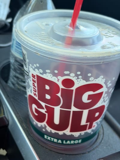 A large, clear plastic cup with a red straw is prominently featured, positioned in a car's cup holder. The cup is branded "SUPER BIG GULP" in bold red lettering, with "EXTRA LARGE" printed in white on a dark green band at the bottom. Numerous small white circular patterns are scattered across the cup, creating the impression of fizz or bubbles from a carbonated drink.
A transparent plastic lid covers the cup, through which a red plastic straw is inserted. Text embossed on the lid is partially visible, including "BIG GULP," and what appears to be "DIET" and "PEPPER," suggesting the beverage is a Diet Dr. Pepper. The liquid level inside the cup is not clearly discernible, but it appears mostly full.
The setting is inside a car, with parts of the dashboard or console visible around the cup holder, rendered in a dark grey or black material. Natural light illuminates the scene, indicating it is daytime. The cup holder and surrounding car interior show some signs of use or dust. The image captures a common sight of a convenience store drink, likely from 7-Eleven (known for Big Gulps), being transported or consumed in a vehicle in the United States.