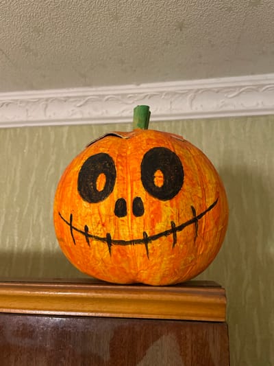 This is a close-up, low-angle shot of a decorative pumpkin that has been painted with a jack-o'-lantern face. The pumpkin is orange with black painted features: two large, round eyes with holes in the center, a small nose, and a mouth that appears to be stitched shut with a vertical line and horizontal dashes. A green stem protrudes from the top of the pumpkin. The pumpkin is positioned to the left of a wooden frame that is leaning against a wall. The wall has a textured, light green wallpaper. To the left of the pumpkin, there is a white decorative trim with a relief pattern. The lighting suggests it is daytime, possibly indoors with artificial light. The overall mood is festive and hints at a Halloween decoration.