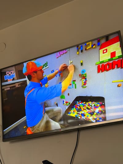 A person wearing an orange and blue hat and a blue shirt is shown on a television screen. The person is building something with Lego blocks, which are spread out on a gray surface. There is also a black container filled with Lego blocks. In the bottom left corner of the screen, there is a logo that says "Blippi". The television is mounted on a wall. The city of Rain, Germany is not visible in the image.