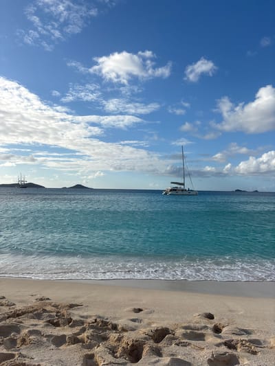 The image depicts a coastal scene in Olde Yard Village, British Virgin Islands, captured from a low vantage point on a sandy beach. The sky, occupying the upper two-thirds of the frame, is bright blue with scattered white cumulus clouds. The horizon line bisects the middle ground, separating the sky from the ocean. The ocean displays a gradient from light turquoise near the shore to deeper blue further out, with small, white-crested waves breaking gently onto the sandy beach.
In the immediate foreground, light-colored sand covers the bottom third of the image, showing numerous distinct footprints and disturbed areas. In the mid-ground, a white catamaran with a single mast is positioned on the water, slightly right of the frame's center. Two small, indistinct human figures are visible on the catamaran's deck; its sails are furled. In the distant background, to the left, a large, multi-masted tall ship with a dark hull is visible near the horizon, partially obscured by a low-lying, dark island. Several other small islands are present along the distant horizon. The scene is illuminated by natural daylight. No specific interactions between individuals or defined actions are clearly discernible.