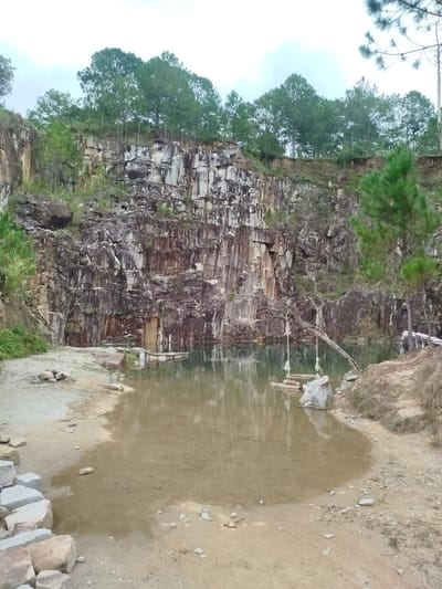 This image depicts a natural quarry in Lán Tranh, Vietnam, transformed into a serene waterhole surrounded by rugged, layered rock formations. Lush green trees, primarily pine, line the top of the quarry, contrasting with the earthy tones of the rock face. A calm, shallow body of water reflects the overcast sky and the surrounding environment. In the foreground, the sandy bank is scattered with rocks of various sizes, some forming a rough path. Further out in the water, a simple wooden raft or platform is visible, anchored by ropes to a tree branch that extends over the water. A swing, also suspended from a tree branch by ropes, is positioned above the water near the raft. The overall scene suggests a tranquil, somewhat undeveloped natural site that has been adapted for leisure. There are no people or animals visible in the image.