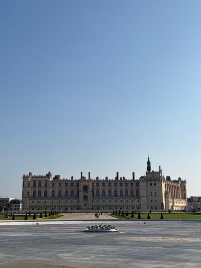 The image displays an exterior, wide-angle view of the Château de Saint-Germain-en-Laye in Saint-Germain-en-Laye, France, under a clear, bright blue sky. The multi-story château, constructed of light-colored stone, features an ornate roofline with multiple chimneys, decorative elements, and numerous arched windows across its facade. A prominent dark spire with a weather vane rises from the rightmost section of the building.
In the foreground, a large, flat, light-grey paved area, resembling a dry fountain basin, is visible. Centered within this area is a circular, dark-colored fountain sculpture depicting multiple figures. Between this foreground area and the château, a wide plaza contains landscaped sections of manicured green lawn, interspersed with rows of evenly spaced conical evergreen shrubs. Approximately ten human figures are observed walking on paved pathways within this plaza, appearing distant and indistinct.
To the far left of the château, white-fronted buildings of an adjacent urban area are partially visible. The upper two-thirds of the image consists solely of a cloudless, light blue sky. A small, dark, elongated object, consistent with an aircraft, is visible in the upper-left quadrant of the sky. The scene is illuminated by natural sunlight from the right, indicating daytime conditions.