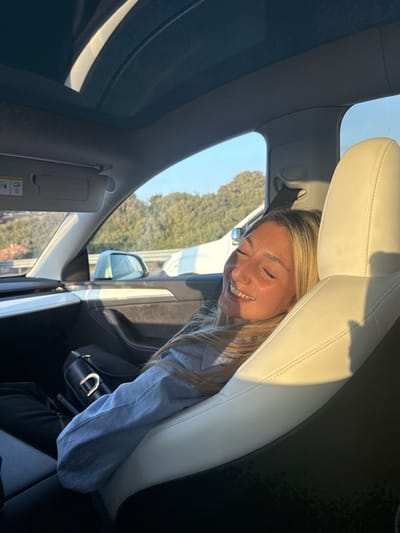The scene depicts the interior of a vehicle in the city of Jaljulia, Israel. A female individual with light-colored hair, wearing a light blue long-sleeved shirt and dark pants, is seated in the front passenger seat. She is observed smiling with her eyes closed, her head tilted slightly back and resting against the light-colored leather headrest. Direct sunlight illuminates her face and the passenger seat. A small metallic nose ring is visible on her right nostril.

The vehicle interior features a light-colored leather passenger seat and dark dashboard/door trim. A black handbag with a silver buckle rests on the floorboard near the passenger's feet. Above, a panoramic glass roof is visible, along with a front sun visor displaying an airbag warning label. Through the passenger window, a clear blue sky is visible above a landscape of green trees and bushes on an incline. A section of a light-colored vehicle is partially visible outside the window, adjacent to the described car. The lighting is natural and bright, indicative of daylight.