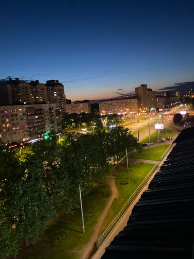 The image depicts a nighttime view in Vaganovo Massiv, Russia. Tall apartment buildings with illuminated windows are prominent on the left, suggesting residential areas. A multi-lane road with streetlights and moving vehicles runs along the center. A green area with trees, a pathway, and lampposts occupies the right side of the image. The sky transitions from blue to dusk in the upper left corner. A dark structure with a white guardrail borders the right edge of the frame, likely a building or bridge.