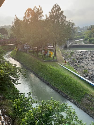 The image depicts a scenic view of a river flowing through a park-like setting, with a playground visible on the grassy bank. The river has a tranquil flow, its surface reflecting the soft, natural light. The banks of the river are lined with stones and lush green grass, suggesting a well-maintained area.
A small playground with a yellow slide and colorful climbing structures is situated under the shade of tall trees, inviting recreational activity. Nearby, there are picnic tables and umbrellas, indicating a space for relaxation and gatherings. The presence of a pedestrian bridge suggests accessibility and connectivity within the area.
In the background, a rocky riverbed is visible, with a dam structure further upstream. The landscape is dotted with trees and vegetation, typical of a tropical or subtropical environment. The sky is overcast with a hazy glow, hinting at either early morning or late afternoon light.
This scene is located in Krasak, Indonesia, a region known for its natural beauty and recreational spots. The image captures a harmonious blend of nature and human-made amenities, creating an inviting atmosphere for visitors.