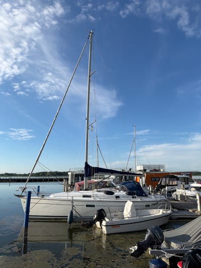 The image shows several sailboats docked at a harbor. The boats are white with blue accents and have tall masts. Some of the boats have outboard motors. The water is calm and reflects the blue sky, which is dotted with white clouds. The sun appears to be shining, suggesting it is daytime. The scene is set in a harbor with a wooden dock and a shoreline with trees in the distance. The overall atmosphere is peaceful and serene.
Visible text on the boats includes "TERHI 450", "BENETEAU", "Diega and", and "YAMAHA". The location context is Ribnitz-Damgarten, Germany. There are no people visible in the image. The activity taking place is the docking of sailboats, suggesting a marina or harbor setting. The time of day is likely daytime, given the bright sunlight and blue sky. The weather appears to be fair.