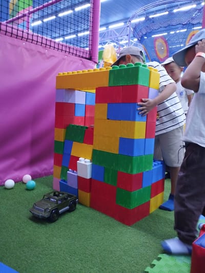 In Talas City, Kyrgyzstan, a group of children are playing in an indoor playground. A large structure built from colorful, oversized building blocks dominates the foreground. The blocks are red, yellow, blue, green, and purple. A toy military jeep is parked on the green carpeted floor in front of the block structure. Several small spherical toys, white, pink, and blue, are scattered near the jeep. Three children are visible, two boys and one whose gender is unclear. One boy, wearing a grey cap and a white and black striped t-shirt, is holding a stack of blocks. Another boy, wearing a blue cap and a white t-shirt, is partially obscured behind the block structure. A third child, seen from the legs up, is wearing a white t-shirt and dark pants, and appears to be standing near the block structure. The background reveals the indoor playground's ceiling with lights and netting.