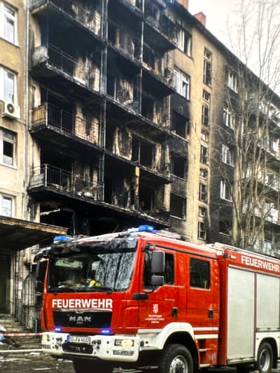 A bright red fire truck with "FEUERWEHR" written on the side is parked in front of a heavily damaged apartment building. The building shows extensive fire damage, with blackened balconies, windows, and facade. Some windows are broken, and debris is visible. The fire truck's lights are illuminated, and its license plate reads "DD-FW 4032". The ground in front of the truck appears wet and possibly covered in snow or ice. The overall scene suggests a recent fire incident in Neubrandenburg, Germany.