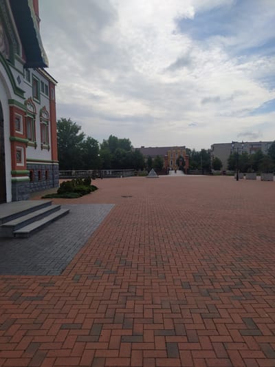 The image shows a view of a town square in Gusev, Russia. In the foreground, there is a paved area with red and dark gray bricks laid in a herringbone pattern. To the left, a building with white, red, and green ornamentation is visible, with a gray stone base and a set of concrete stairs leading up to it. Beyond the square, there are trees, buildings of various architectural styles, and streetlights. The sky is cloudy with patches of blue. There are no people visible in the immediate foreground or mid-ground. The scene appears to be a public space with a mix of historical and modern architecture.