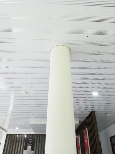 The image captures an interior view within a commercial establishment in Pachmarhi, India. A prominent cream-colored cylindrical column stands centrally, extending vertically from an unseen base to a white paneled ceiling. The ceiling features parallel grey lines and a subtle textured wave pattern, equipped with multiple recessed circular light fixtures, one of which is brightly illuminated. A dark, soiled ring is evident at the column's apex where it meets the ceiling. Partial views of dark brown slatted partitions are visible to the lower left and right of the column. A black promotional poster, depicting a glass of a white beverage and text "Jus Fresh," is affixed to the left partition. Two additional vertical red-orange posters are partially visible on the right partition. A dark framed opening, possibly a window or doorway, is visible in the far right background. No individuals or dynamic activities are observed.