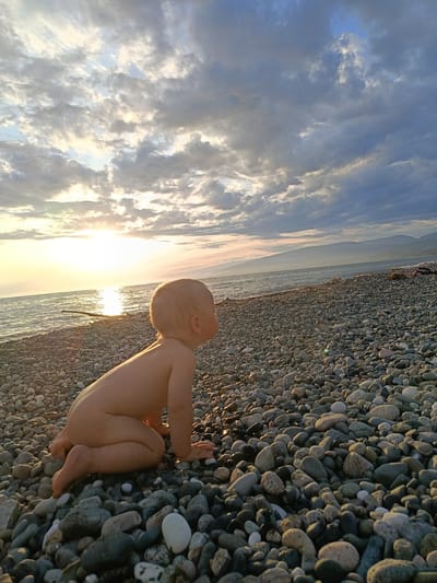 The image depicts a baby, from the waist down, sitting on a pebble beach. The baby is facing away from the camera, looking towards the sea. The sun is setting over the water, casting a warm glow. In the background, mountains can be seen. The overall scene suggests a peaceful and serene setting. The location is Gulrypsh, Abkhazia.