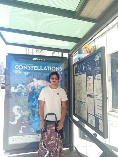 A young man with dark hair, sunglasses, and a light-colored t-shirt stands at a bus stop in Nancy, France. He is holding a patterned shopping trolley with a burgundy background and white text featuring various city names. Behind him is a bus stop shelter with a green, meshed roof. To his left is a large poster advertising the "FESTIVAL INTERNATIONAL CONSTELLATIONS" from June 25 to September 5, 2026, with the JCDecaux logo at the top. To his right, a large information board displays maps and details about local transportation services, including a QR code. The bus stop is situated on a street with buildings visible in the background.