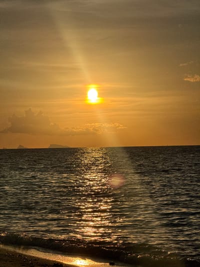 This media file depicts a sunset at Baan Tai, Thailand. The image is split vertically, with the left side showing the sun setting behind a hazy, golden sky. Streaks of light emanate from the sun, creating a diffused glow that illuminates wispy clouds. On the right side of the image, the ocean is visible, with the setting sun casting shimmering reflections on the rippling water. The waves appear dark, contrasted by the bright glints of sunlight. The edge of the land, possibly a beach, is visible on the far right, with wet sand reflecting the golden light. The overall mood is serene and peaceful, capturing the beauty of a tropical sunset. The time of day is clearly sunset, and the weather appears clear with some atmospheric haze. There is no visible text in the image.