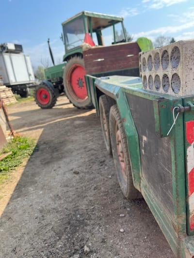 The image depicts an outdoor scene in Krško, Slovenia, likely a farmyard or industrial area, under clear daylight conditions. In the immediate foreground, to the right, is a green, multi-axle flatbed trailer with red wheel hubs and black tires. The trailer's side panels display wear, scratches, and dirt. Stacked on the trailer bed are at least two light-colored, rectangular concrete blocks, each featuring two rows of six circular perforations. A white metal hairpin cotter pin is visible on a latch mechanism at the rear right side of the trailer, which also has a red and white diagonal reflective safety stripe.
In the midground, to the left of the trailer and out of focus, is a green agricultural tractor with large red wheels and a green cab enclosure. A red plastic object is positioned on the tractor's engine hood area. In the far background, further to the left and also out of focus, a portion of a white truck or semi-trailer is visible. The ground is an unpaved surface consisting of packed dirt and gravel, with various small stones and irregularities. Sparse, bare tree branches are visible against a clear blue sky in the upper background. No individuals are present in the scene.