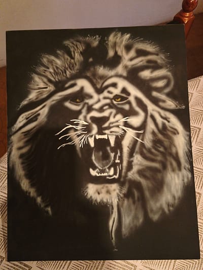 A painting of a roaring lion with a black background is on a table with a patterned cloth in Sant Esteve Sesrovires, Spain. The lion has a large mane and its mouth is open revealing its teeth. Its eyes are yellow. The painting appears to be on a canvas frame. A small portion of a wooden chair is visible in the background.